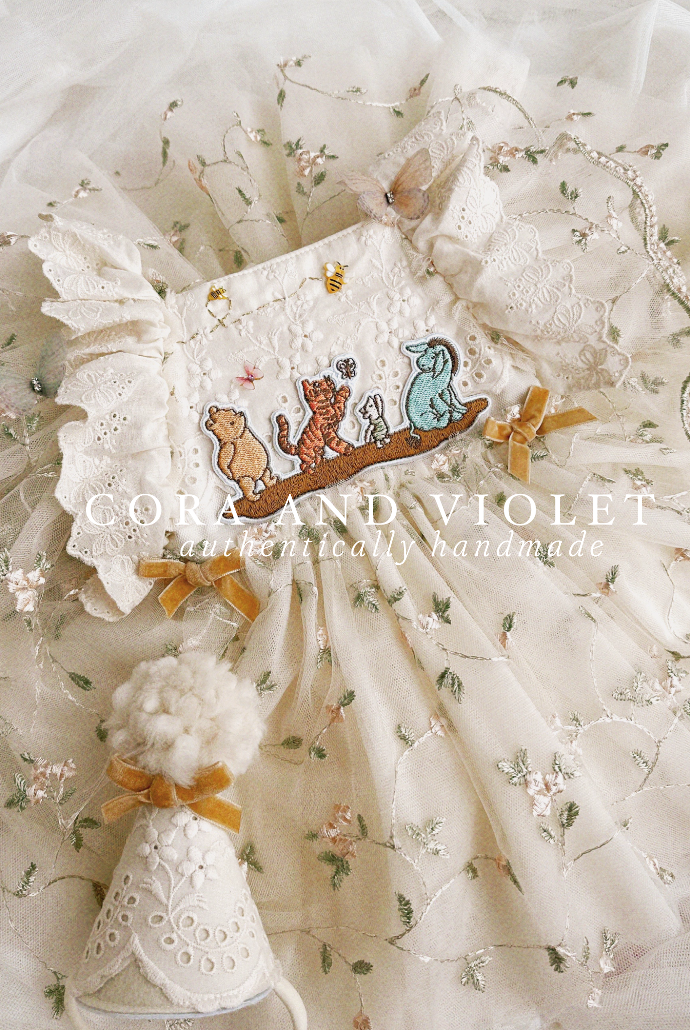 Close-up of artisan-stitched rosebuds and hand-applied butterflies on a luxury classic pooh milestone gown. Storybook Set featuring the Winnie dress, a rose party hat, and a honey bee birthday wand for a first birthday celebration.