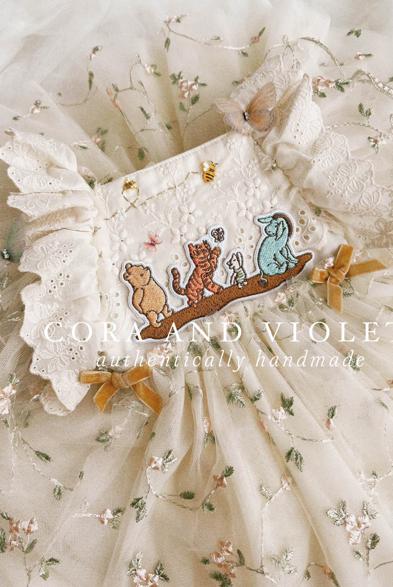 Close-up of artisan-stitched rosebuds and hand-applied butterflies on a luxury classic pooh milestone gown. Storybook Set featuring the Winnie dress, a rose party hat, and a honey bee birthday wand for a first birthday celebration.