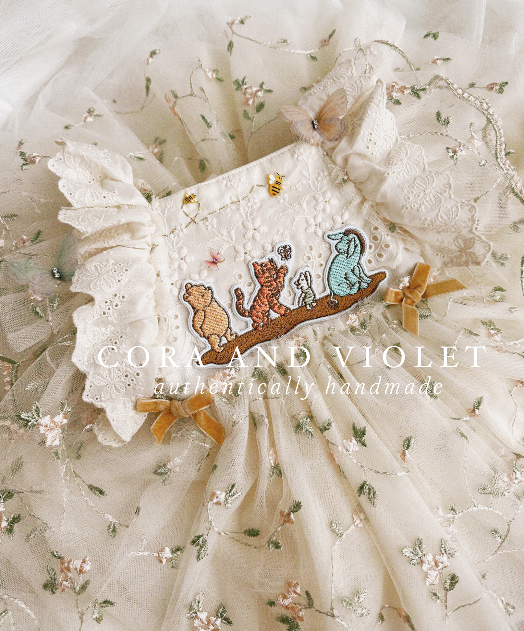 Close-up of artisan-stitched rosebuds and hand-applied butterflies on a luxury classic pooh milestone gown. Storybook Set featuring the Winnie dress, a rose party hat, and a honey bee birthday wand for a first birthday celebration.