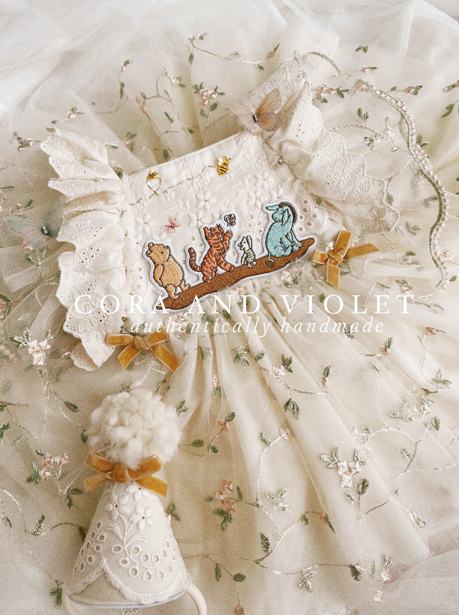 Close-up of artisan-stitched rosebuds and hand-applied butterflies on a luxury classic pooh milestone gown. Storybook Set featuring the Winnie dress, a rose party hat, and a honey bee birthday wand for a first birthday celebration.